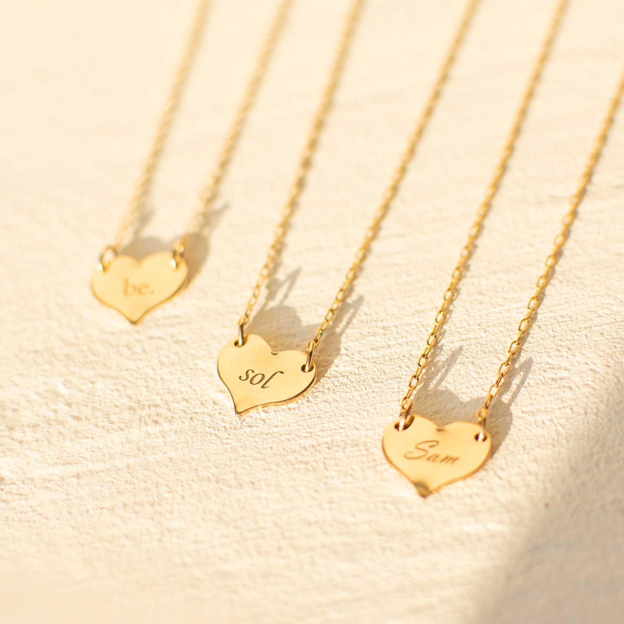 Three gold heart pendants are laying against a white texture board. The left heart is out of focus and has be with a period engraved in the ol' serif font. The middle heart is in focus and has sol engraved in the corsiva font. The right heart has Sam engraved in the script font and is out of focus. 