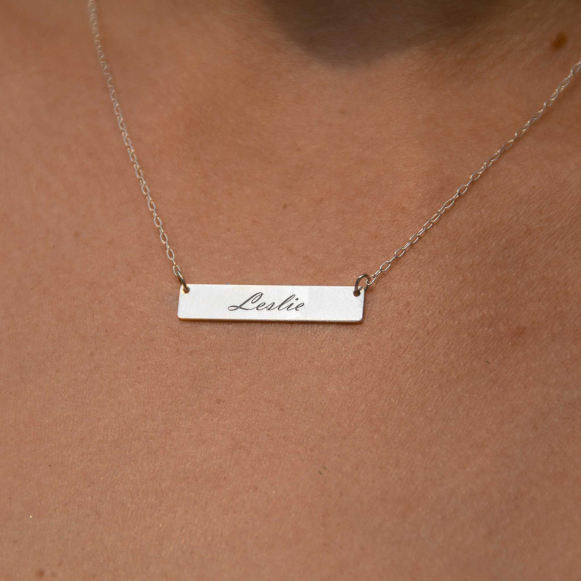 Up close of the rectangle bar necklace worn with Leslie engraved in the script font. 