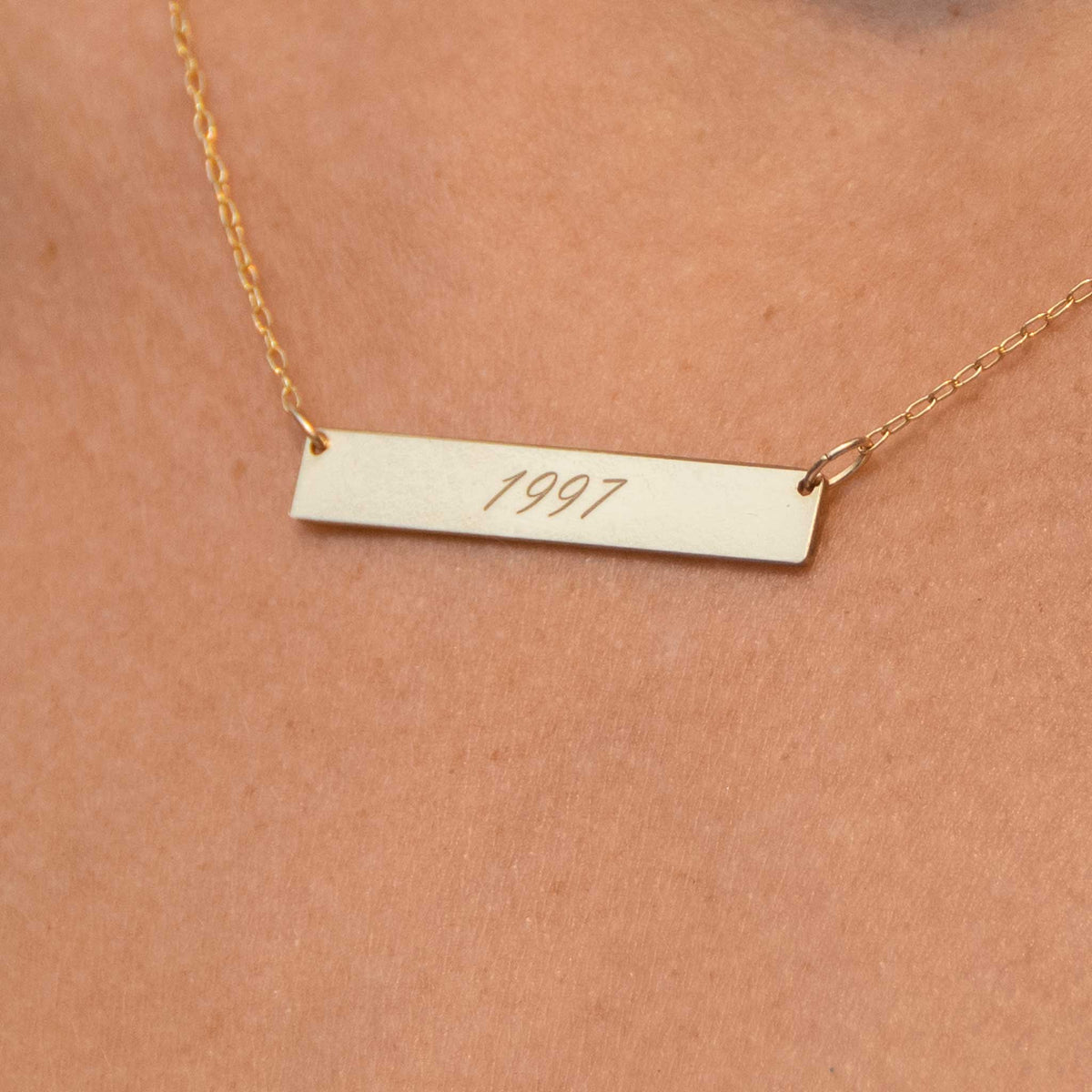 A very close up of the rectangle bar necklace in gold color with 1997 engraved in script font. 