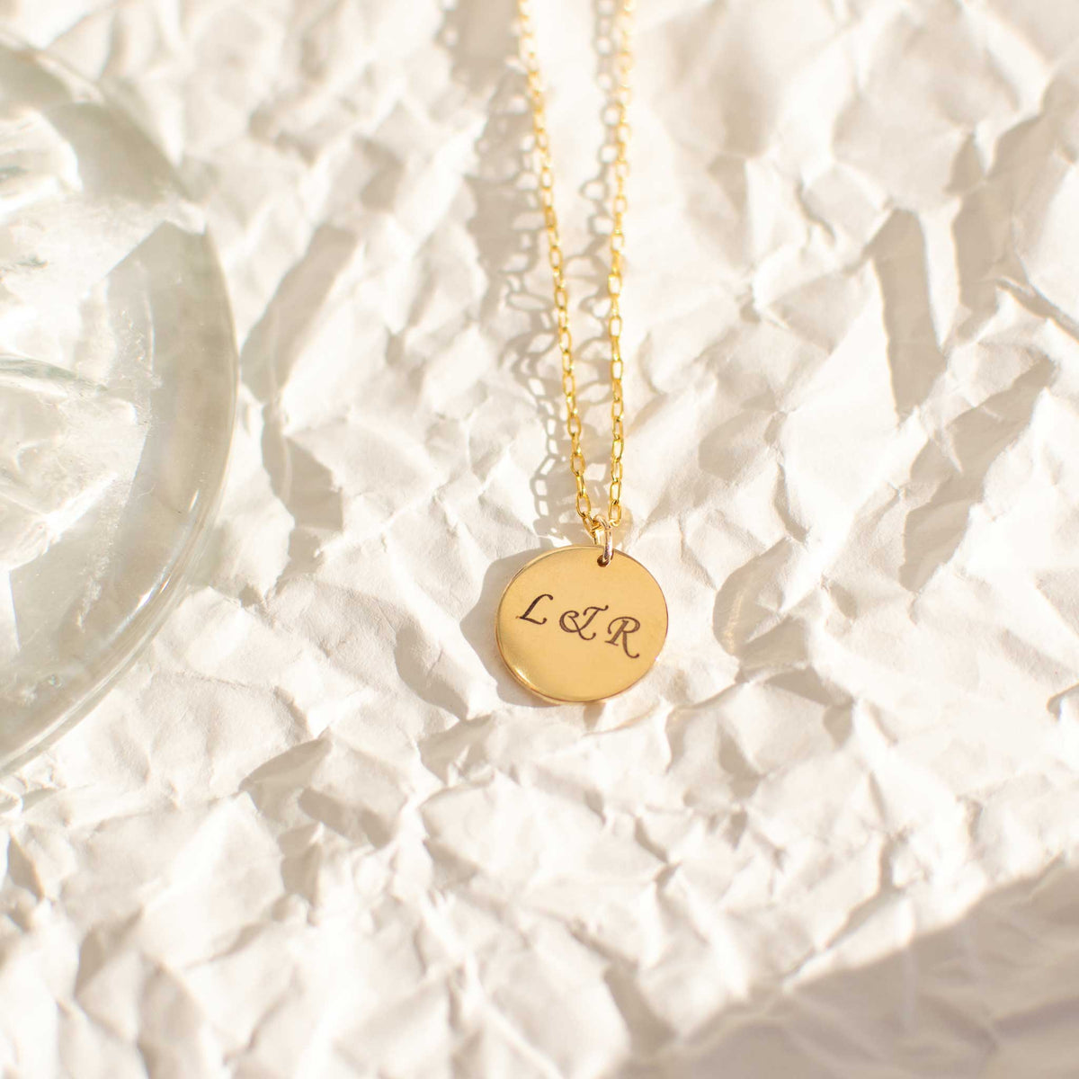 The 13mm gold circle pendant with L &amp; R engraved. The disk is laying on crumpled white paper with direct harsh lighting, and the bottom of a wine glass is on its left. 