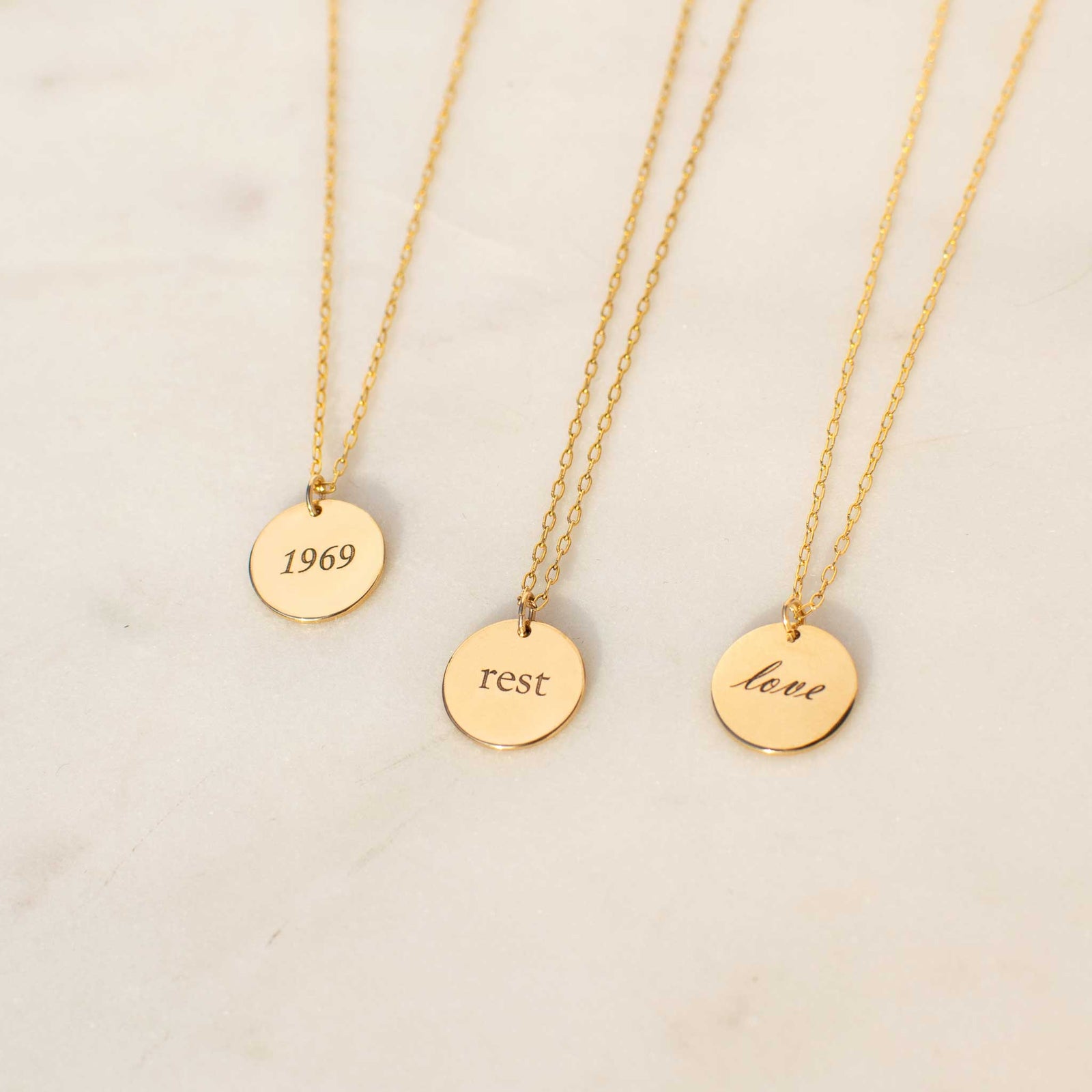 Three  13mm size circle disk pendants are laid down on a white marble background. The left circle has 1969 engraved into it in the corsiva font, the middle circle has rest engraved in the ol' serif font, and the right circle has love engraved with the script font.