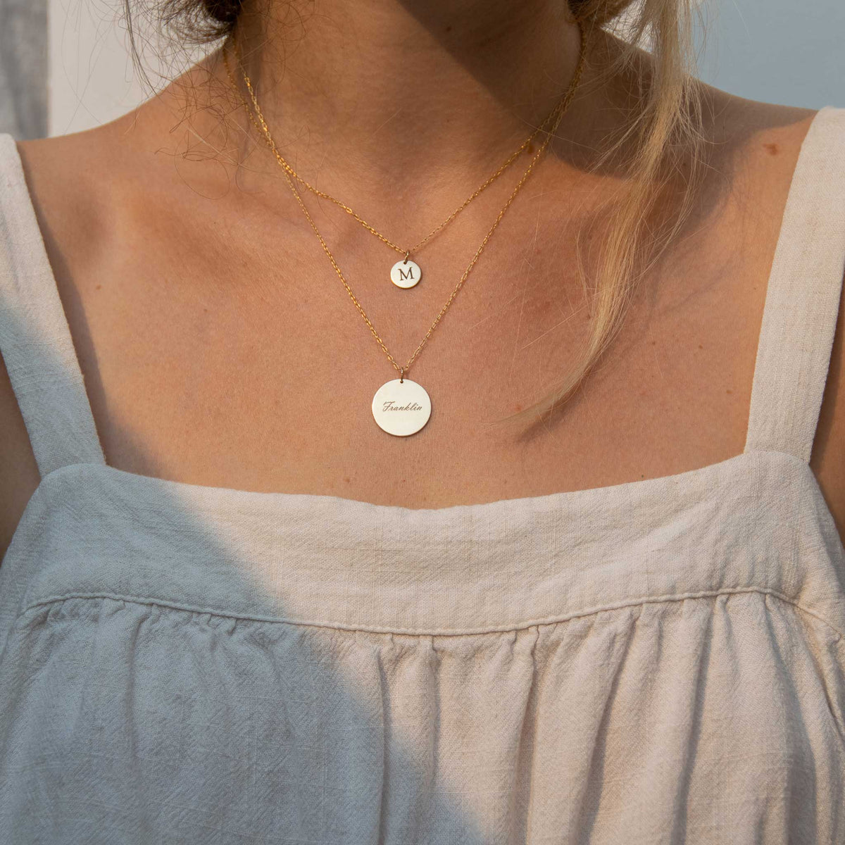 A woman wearing a small 10mm circle necklace with M engraved, and the 19mm circle necklace layered below on a longer chain with Franklin engraved. 