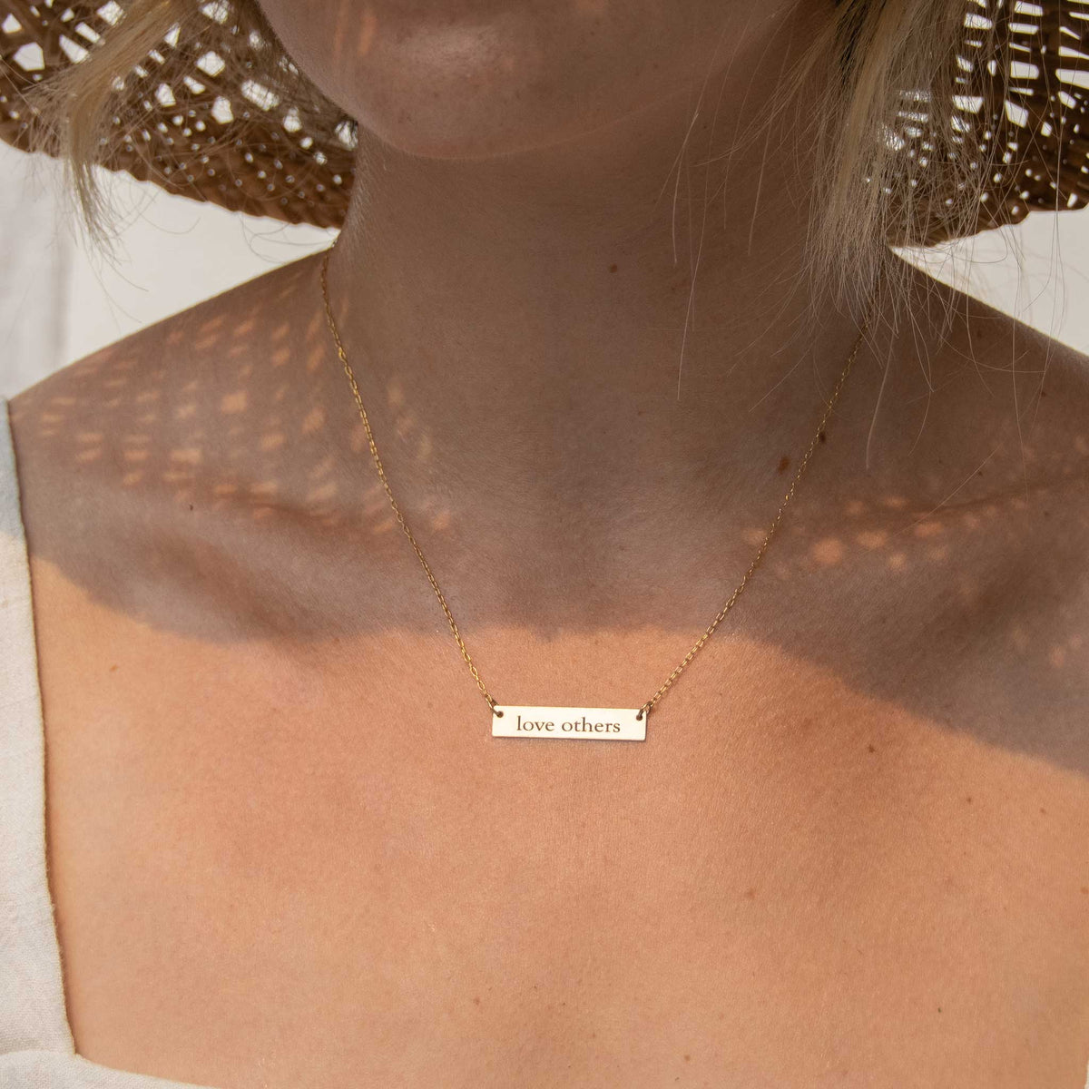 a woman wearing the 1.25 bar necklace with love other engraved with shadows from her woven hat casting geometric shadows onto her shoulders. 