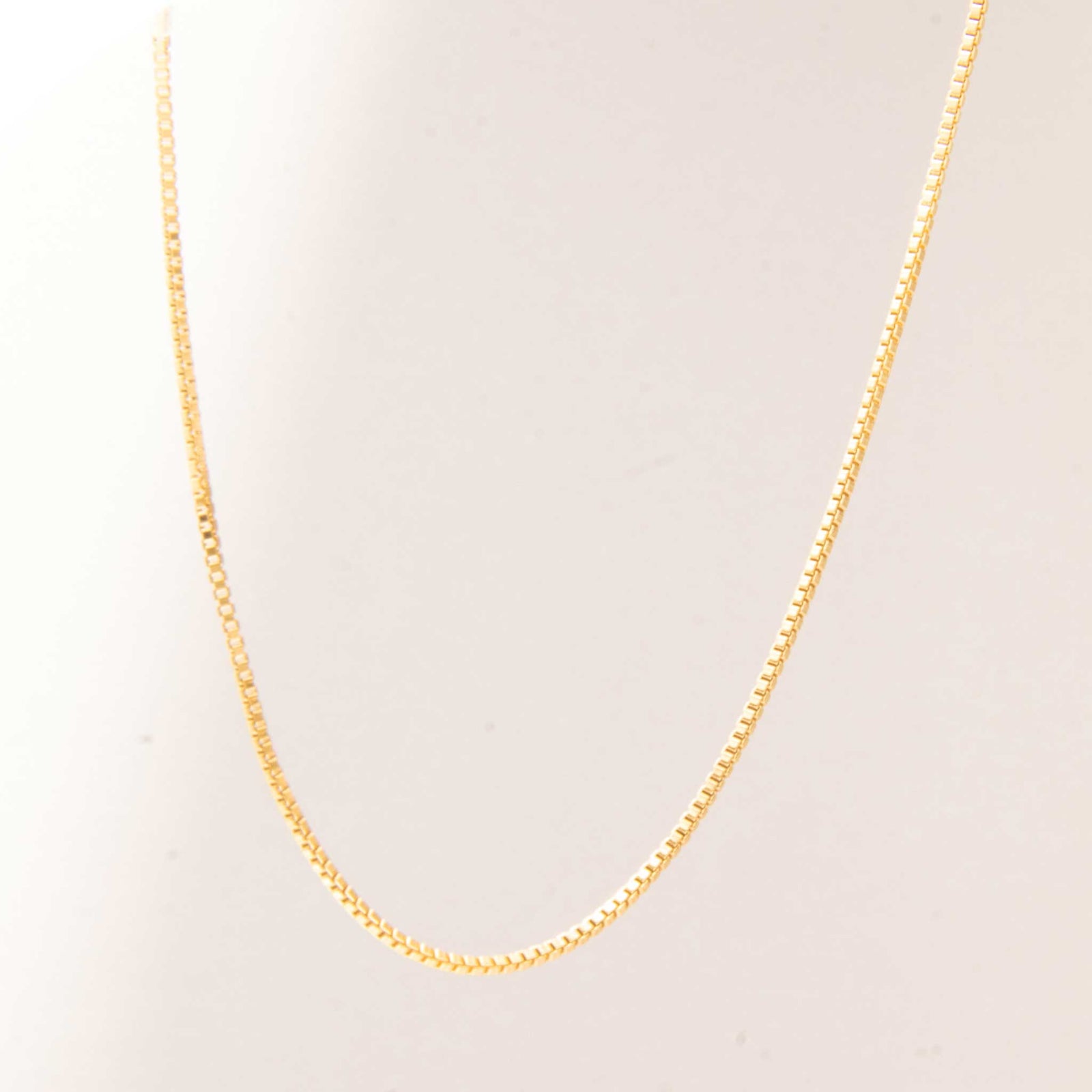 A close up of gold box chain necklace hanging from above with a white color background.