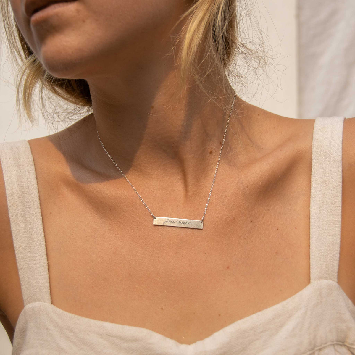 Woman looking to the left wearing a silver rectangle bar necklace with josie raina engraved in the script font. 