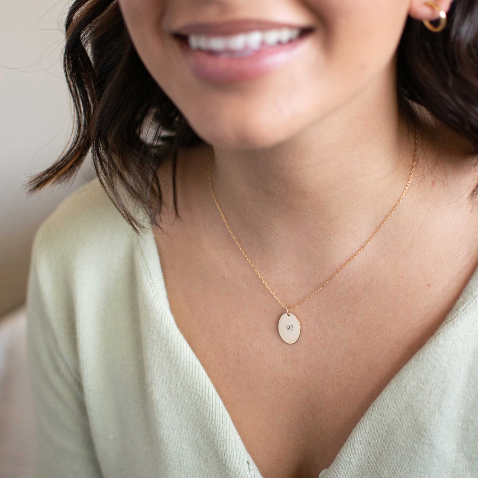 Woman wearing the gold oval pendant with '97 engraved in the ol'serif font while also wearing a v neck blouse. 