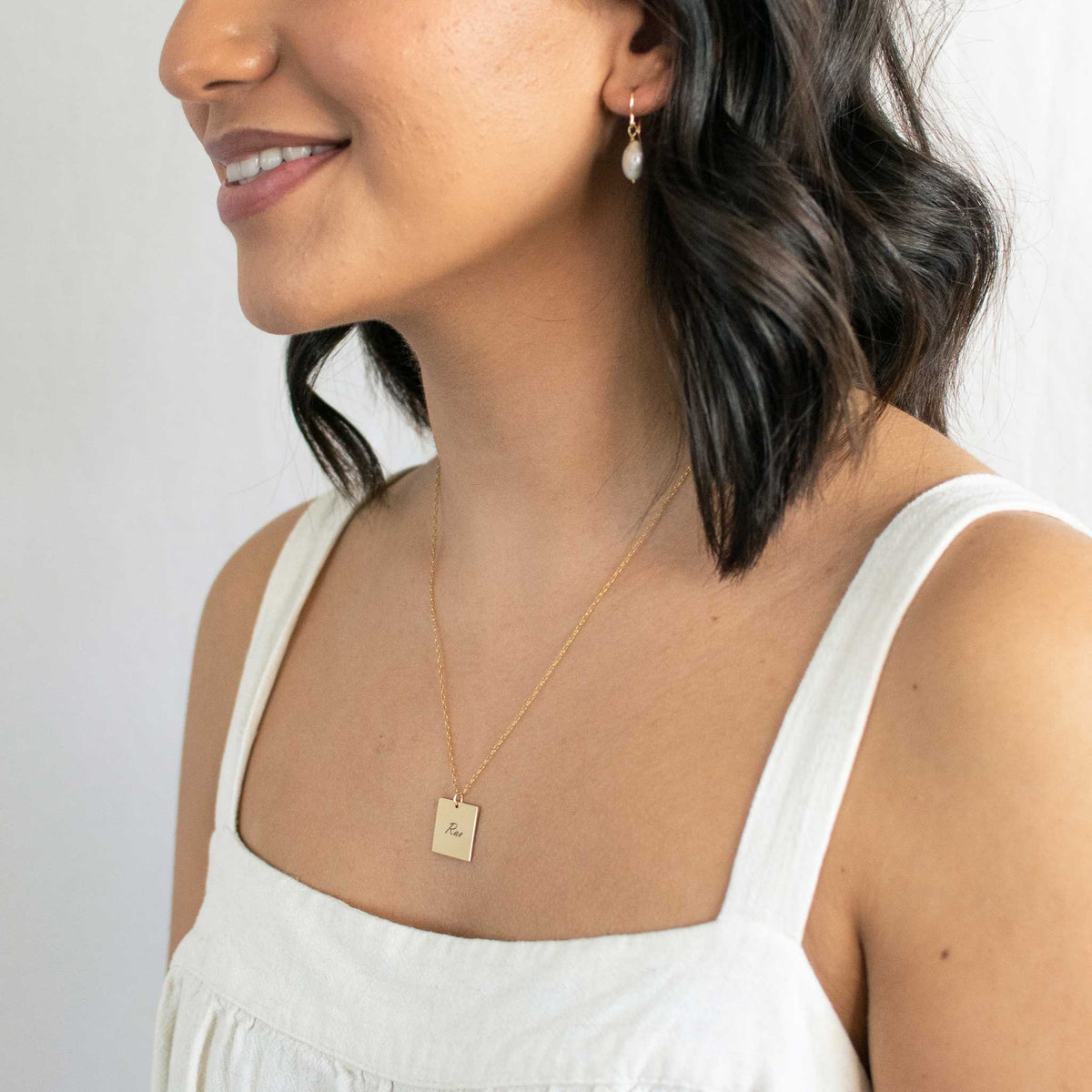 Woman is smiling while wearing a gold square pendant with Rae engraved in the font script. 